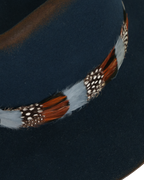 Close-up view of a navy wide-brim fedora with a teardrop crown, multicolor feather hat band in rust, white, and speckled tones.
